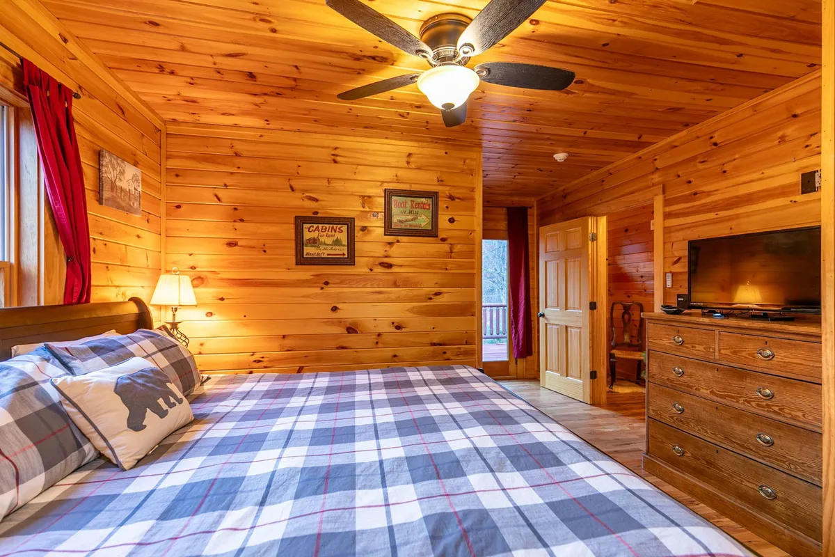 A view of a bedroom featuring plaid bedding, wooden walls, a ceiling fan, bedside lamps, and a detailed wooden dresser. The ambiance feels relaxed and inviting, perfect for restful nights after a day of adventure.