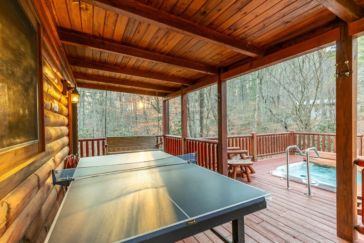 This image features an outdoor wooden deck surrounded by trees, showcasing a ping pong table and a hot tub. Several people are engaged in playful and relaxing activities, such as playing ping pong, lounging in the hot tub, and socializing. The setting is lively and inviting, with hints of a sunset in the background.