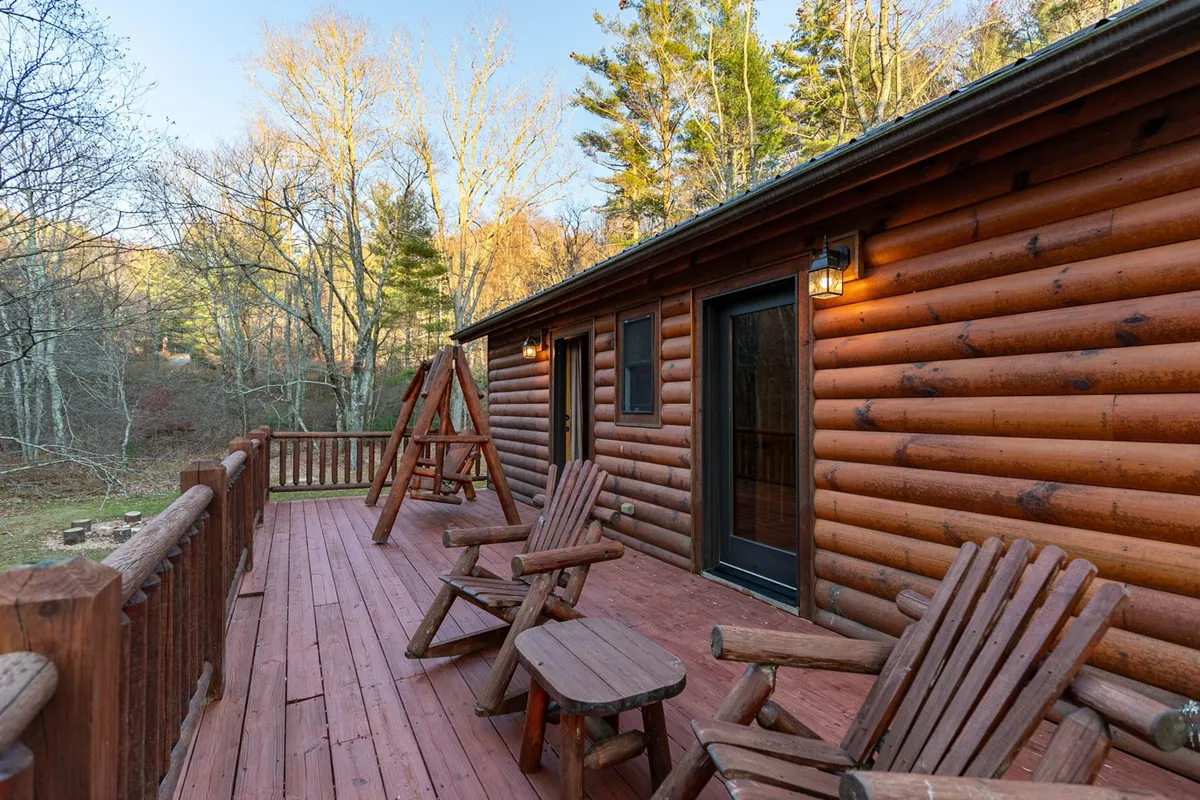 A cozy log cabin exterior featuring a spacious wooden deck outfitted with Adirondack chairs and a swing set. The setting is tranquil, with inviting spaces for group relaxation, enjoying nature, and unwinding together.