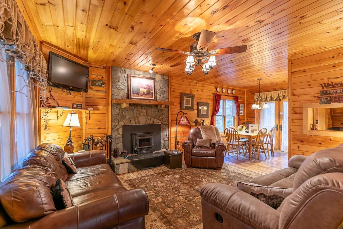 A warm and welcoming living room featuring a stone fireplace, ample leather sofas arranged for conversation, and a sturdy dining table in the background. Sunlight pours in, creating a cozy, inviting atmosphere ideal for family gatherings or quiet evenings by the fire.