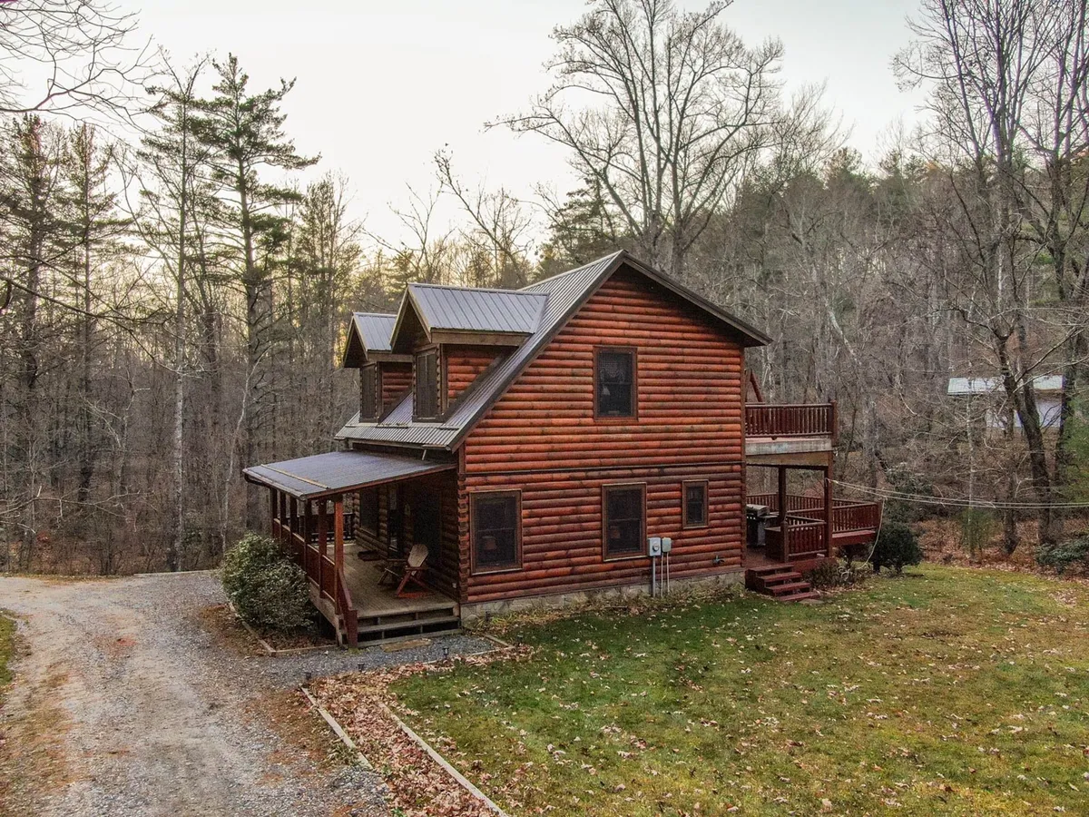 This image features the log cabin exterior with a clear view of the surrounding trees, lawn area, and a gravel driveway leading up to the house. Ample parking space is visible, and the inviting porch hints at a well-managed, welcoming property.