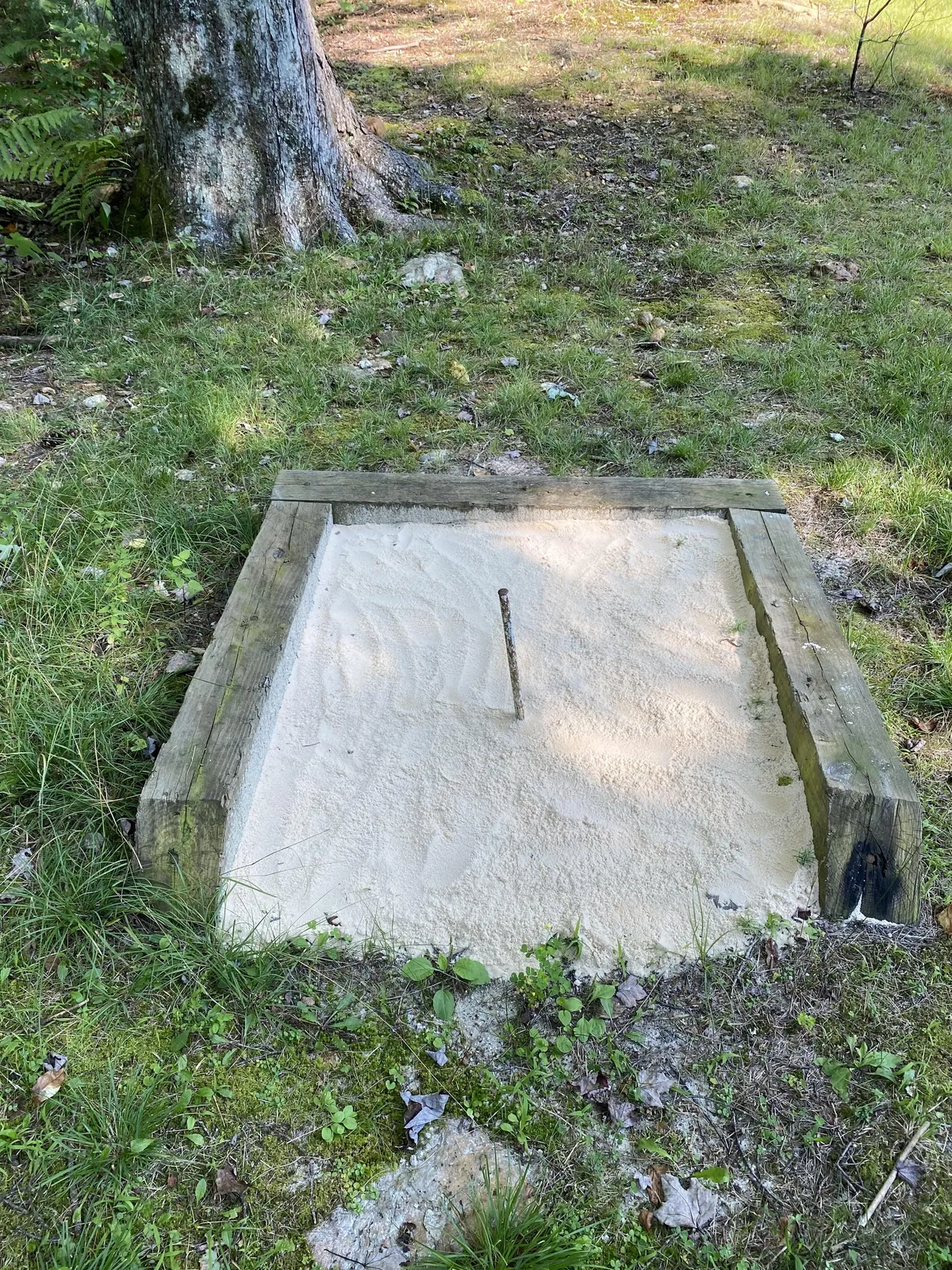 A playful sandbox set amidst green grass, suggesting a spot where children can build sandcastles, explore, and play freely. The simple, cheerful setting hints at family gatherings and child-friendly outdoor fun.
