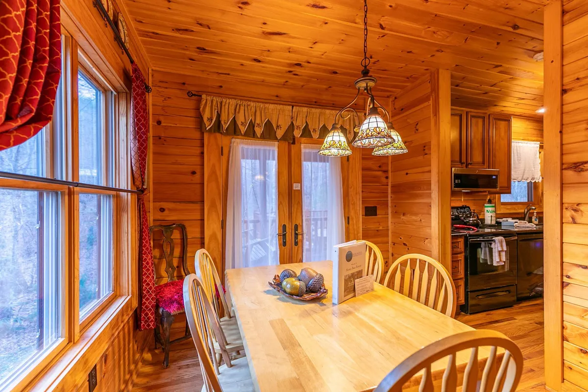 A wooden dining table surrounded by comfortable chairs, with stained glass light fixtures overhead and kitchen appliances in view. The setting is intimate and ready for family dinners or lively gatherings.