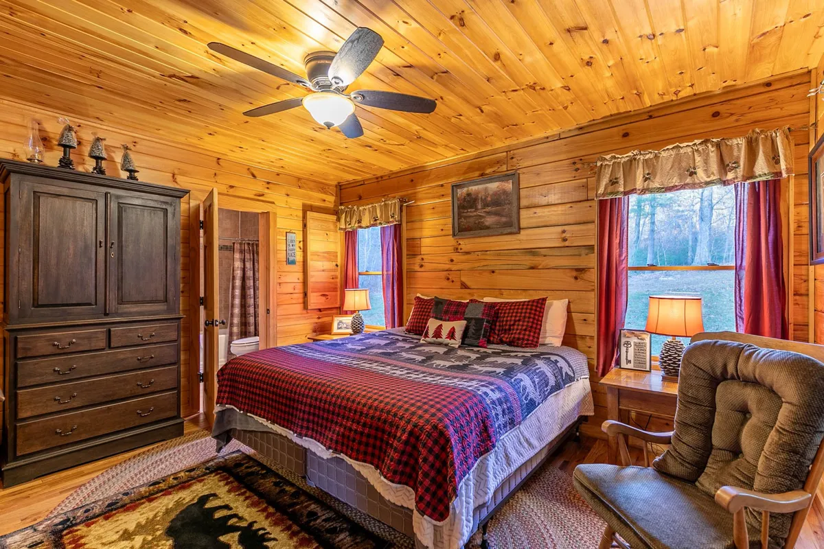 This image showcases a bedroom with a bed adorned in patterned bedding, a wooden dresser, windows letting in natural light, and a cozy rocking chair. The overall setting radiates warmth and comfort, reinforcing the idea of a restful and welcoming retreat.