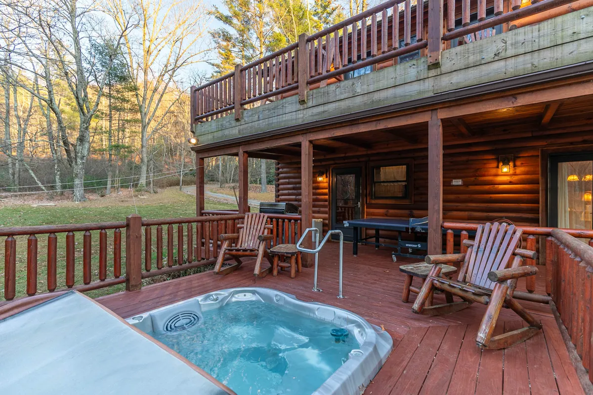 The image highlights a wooden deck featuring a hot tub adjacent to the log cabin structure, set against a picturesque backdrop. The scene is ideal for relaxing in the hot tub or hosting outdoor gatherings at sunset, with mountain scenery visible in the background.