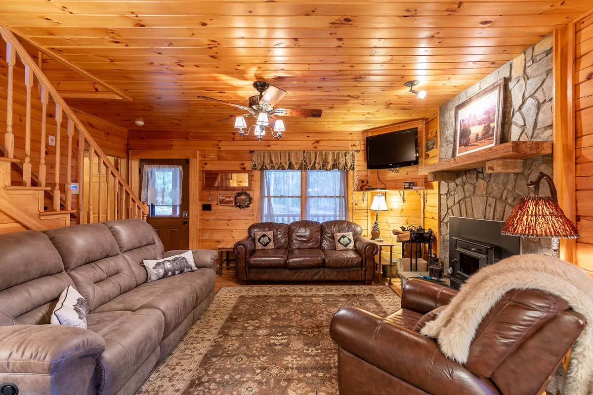 An inviting open space with leather sofas facing a stone fireplace. A wooden staircase and art on the walls add character. The room is filled with natural light, emphasizing a sense of openness and comfort.