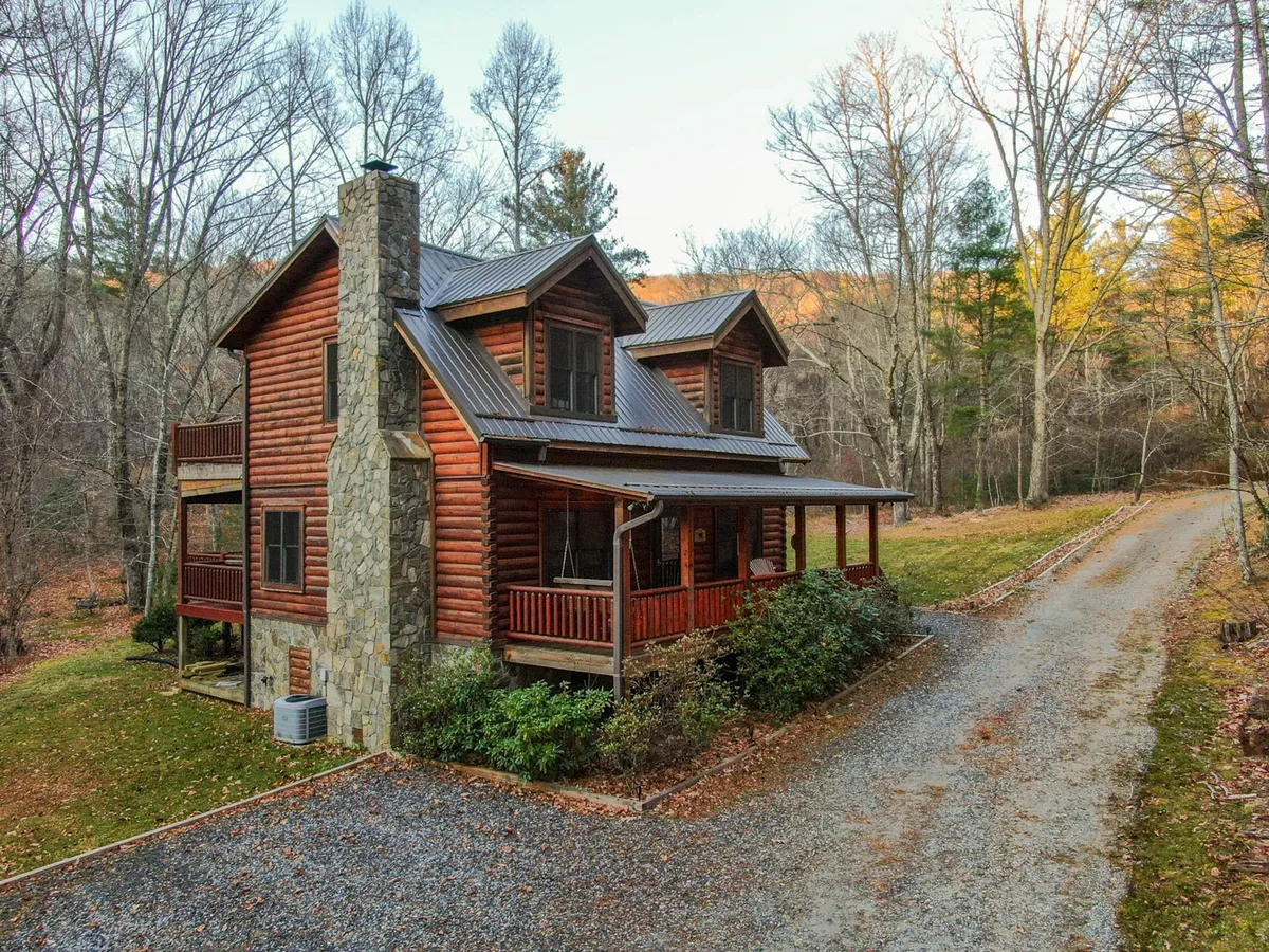 The image features a charming log cabin-style home with a striking stone chimney, nestled among tall, lush trees. The inviting porch overlooks tranquil woods, perfect for catching sunsets, while the peaceful surroundings promise seclusion. The scene evokes warmth and comfort, setting the tone for outdoor gatherings and relaxation.