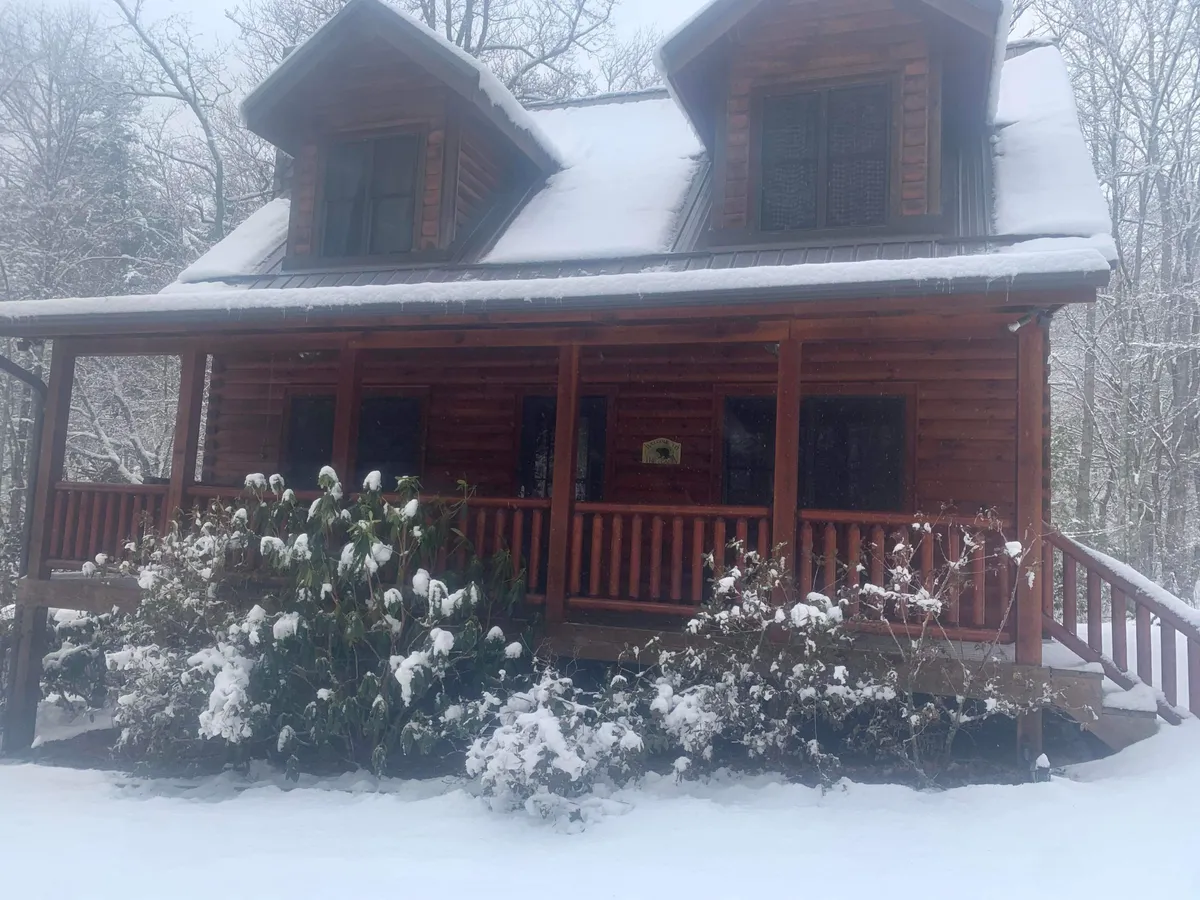 A log cabin with a snow-covered roof and wooden porch, surrounded by trees and snow-dusted plants. The scene highlights the cabin's inviting exterior set against a winter landscape, evoking adventure and the need for suitable vehicles during icy or snowy months.