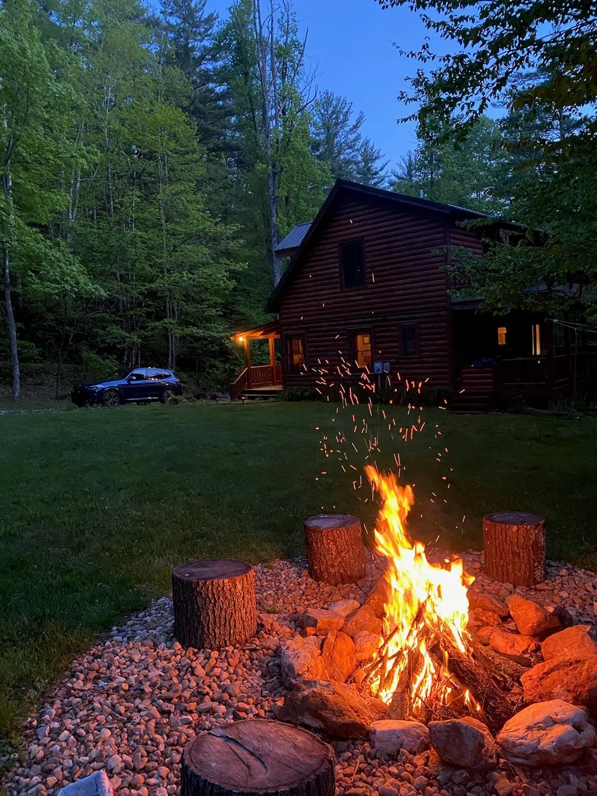A cozy fire pit scene with log seating encircling a lively campfire, set against the backdrop of a cabin and a lush tree line. The setting invites gatherings, storytelling, and the enjoyment of roasted marshmallows under the stars.