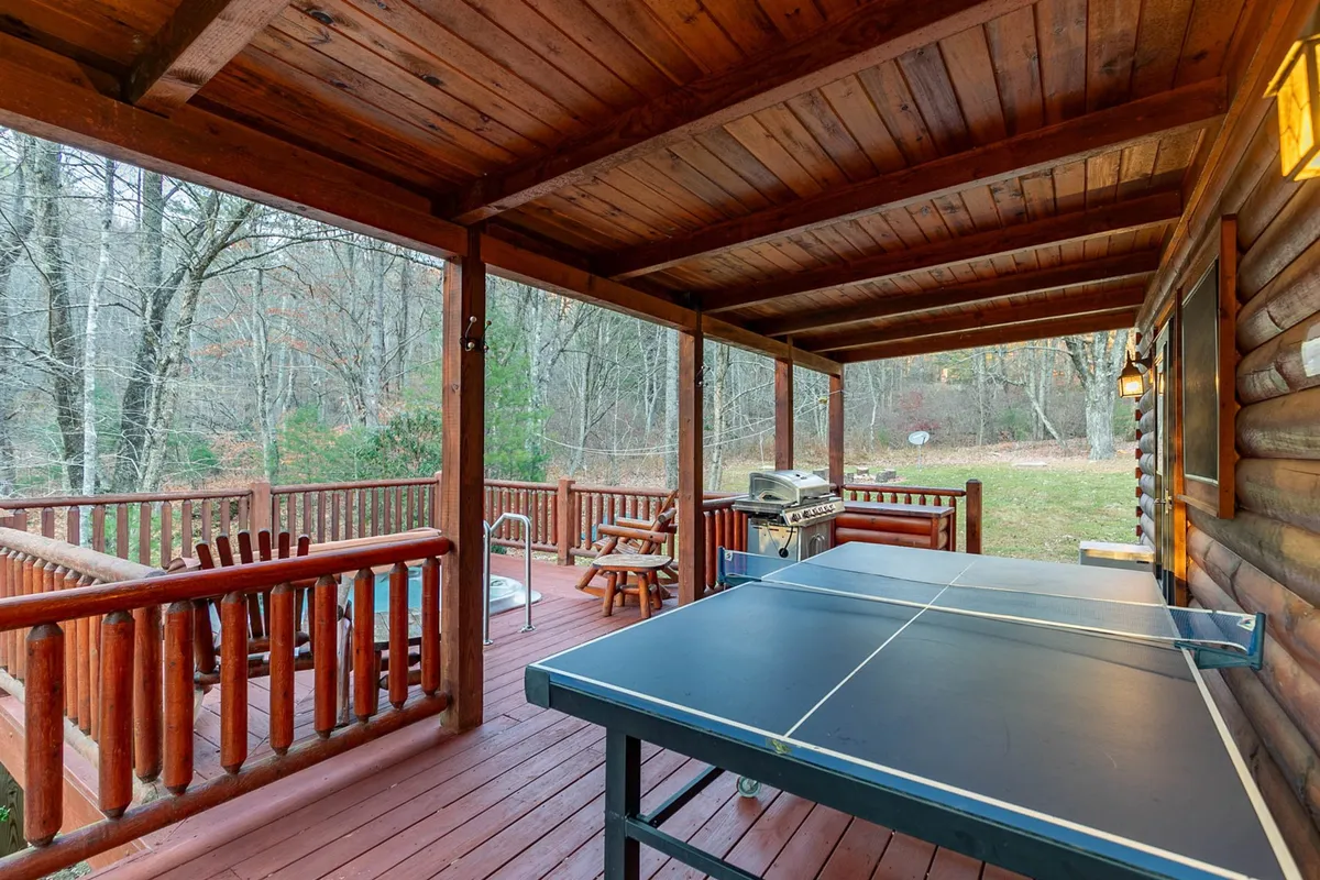 An inviting outdoor deck with a ping pong table, grill, and wooden chairs set against a backdrop of natural scenery. The space is set up for social gatherings, with plenty of room for guests to relax, play games, and dine outdoors.