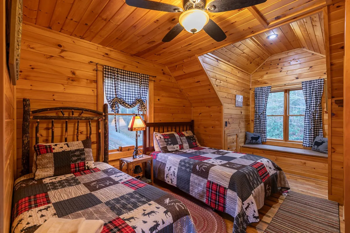 This image features two wooden beds with patchwork quilts, a bedside lamp casting a warm glow, and a window with a peaceful outdoor view. The scene is inviting, offering a sense of shared comfort and quiet relaxation.